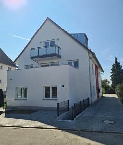 Modernes Wohnhaus mit Satteldach, weißer Fassade und Glasbalkon, Vorgarten und Einfahrt, bei sonnigem Wetter in ruhiger Wohngegend.