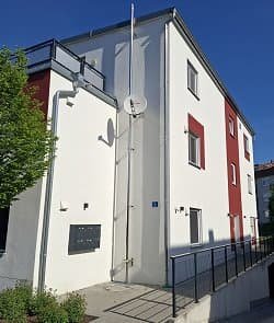 Modernes Mehrfamilienhaus mit weiß-roter Fassade und Satellitenschüssel bei blauem Himmel, aufgenommen aus schrägem Winkel.
